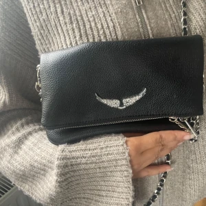 Zadig &Voltaire väska  - Säljer en svart axelväska i skinnimitation med silverfärgad kedja och ett glittrigt emblem framtill. Väskan har dragkedja och är perfekt för att bära det viktigaste. Stilren och enkel att matcha till olika outfits. ( väska är inte äkta)