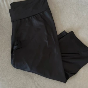 Svarta trekvartsleggings från Lindex - Svarta trekvartsleggings från Lindex med bred midjeresår. Snyggt glansigt material som sitter tight och är perfekta för träning eller chill. Enkel och stilren design utan synliga detaljer.