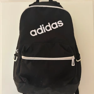 Svart Adidas ryggsäck  - Svart ryggsäck från Adidas med stor vit logga framtill. Väskan har ett stort huvudfack, en framficka med vit dragkedja och vadderade axelremmar. Se sista bild för defekter. Tillverkad i slitstark polyester och perfekt för skola eller träning 🌸🌸