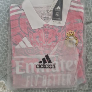 Real Madrid rosa fotbollströja Adidas M/S - Säljer en officiell Real Madrid fotbollströja från Adidas i rosa med ljusgrått drakmönster. Tröjan har vit krage, korta ärmar och klassiska Adidas-ränder på axlarna. Materialet är lätt och andas, perfekt för träning eller match. Emirates Fly Better-logga på bröstet.