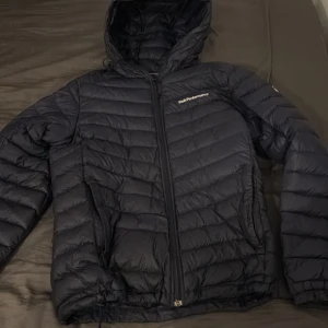 Svart dunjacka Peak Performance - Säljer en svart dunjacka från Peak Performance med huva och dragkedja. Jackan är quiltad och har en stilren design med logga på bröstet och ärmen. Perfekt för kalla dagar och enkel att matcha med det mesta.