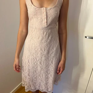 Dammrosa spetsklänning med knappar - Superfin Dammrosa kortklänning i spets med ärmlös design och rundad urringning. Klänningen har dekorativa knappar framtill!