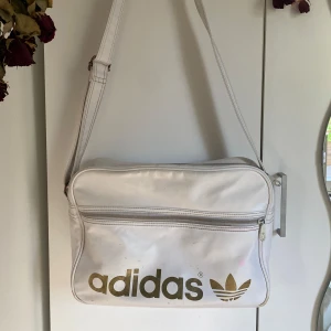 Vit axelremsväska Adidas - OG ADIDAS airliner-väska. Välanvänd, men fortfarande i bra skick. Den är gjord i konstläder. Väskan har justerbar axelrem, dragkedja framtill och rymligt huvudfack. Vissa fläckar går troligtvis bort vid tvätt