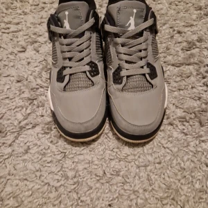 Gråa Nike Air Jordan 4 sneakers - Snygga Nike Air Jordan 4 sneakers i grått med svarta och vita detaljer. Skorna har klassisk snörning, meshpaneler på sidorna och en robust sula. Ikonisk Jumpman-logga på plösen och häl. Perfekta för dig som gillar streetstyle och vill sticka ut.