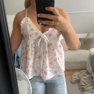 Vit blommig linnetopp med volang - Supersöt vit linnetopp med rosa och gula blommor, volangdetalj vid axeln och knyt framtill. Tunn och luftig med v-ringning och smala axelband, perfekt för varma dagar.