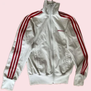 Vit Adidas track jacket XS - Vit Adidas track jacket med klassiska röda ränder längs ärmarna och broderad logga på bröstet. Jackan har hög krage, dragkedja framtill och två fickor. Tillverkad i mjuk bomullsblandning, perfekt för en sportig och retro vibe. Den är mammas gamla från 2000s så den är använd men inga stora tecken på det.