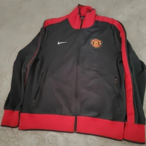 Manchester United Nike track jacket L - Snygg svart och röd Manchester United track jacket från Nike i storlek L. Jackan har dragkedja, ståkrage och klubbens emblem på bröstet samt stor logga på ryggen. Perfekt för fotbollsfans som vill sticka ut med stilren sportig look. Manchester United jätte fin har några  noppor annars är allt bra. har manhister United på ryggen stolekt L
