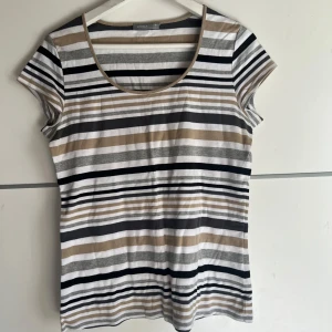 Randig topp från Lindex i beige och svart - Snygg randig topp från Lindex i storlek M. T-shirten har korta ärmar och rund halsringning. Färgerna går i beige, svart, grått och vitt med horisontella ränder. Perfekt för en chill och stilren look.
