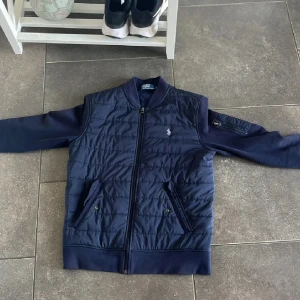 Mörkblå cardigan från Polo Ralph Lauren - Snygg mörkblå cardigan från Polo Ralph Lauren med quiltad front, ribbade muddar och ärmar i mjuk bomullsmix. Jackan har dragkedja, två fickor fram och en extra ficka med dragkedja på ärmen. Klassisk logga broderad på bröstet.