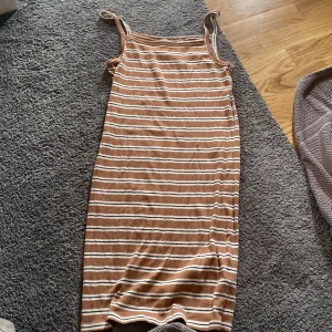 Brun och vit randig klänning H&M - Superfin ribbad långklänning från H&M med smala axelband. Klänningen är brun med vita och mörkbruna horisontella ränder och har en figurnära passform. Perfekt för varma dagar och enkel att styla med sneakers eller sandaler.