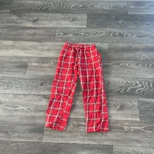 Röda rutiga byxor från 157 - Röda byxor från 157 med klassiskt rutigt mönster i vitt och svart. De har resår i midjan och snörning för extra komfort. Perfekta för chill dagar hemma eller när du vill ha en avslappnad look. Tillverkade i mjukt bomullstyg.