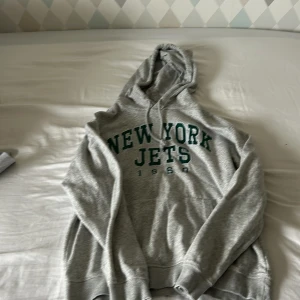 Grå New York Jets hoodie XS - Snygg grå hoodie med New York Jets-tryck i grönt framtill. Klassisk huva med snörning och mjuk insida. Perfekt för dig som gillar sportig stil och vill ha något bekvämt och avslappnat. Passar dig som vill sticka ut med amerikansk college-vibe.