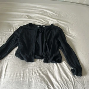 Svart kofta med knyt från lager 157 - Svart kofta från 57/8 i mjukt material. Den har långa ärmar och en söt knytning vid halsen som ger en extra detalj. Perfekt att slänga över en topp för en chill look. Passar dig som gillar enkla men snygga plagg.