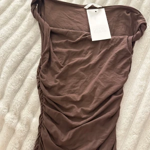 Brun topp från zara - Säljer en brun topp från Zara i mjuk bomull. Tröjan har snygg rynkdetalj längs ena sidan och en enkel, ärmlös design med bred halsringning. Perfekt för dig som gillar minimalistisk och trendig stil. Toppen går inte att köpa längre!