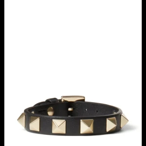Valentino armband - Coolt svart armband i skinn med fyrkantiga guldfärgade nitar runtom. Justerbart spänne för perfekt passform. Perfekt accessoar för att ge din outfit en edgy vibe. Påse och låda följer med