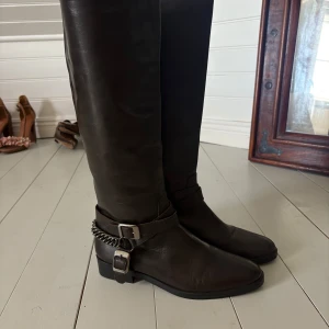 Mörkbruna skinnstövlar med spännen - Snygga mörkbruna stövlar i skinn med coola detaljer som dubbla spännen och en kedja runt ankeln. De har låg klack, så väldigt sköna samtidigt som de är otroligt snygga!! Väldigt sparsamt använda, inga defekter 