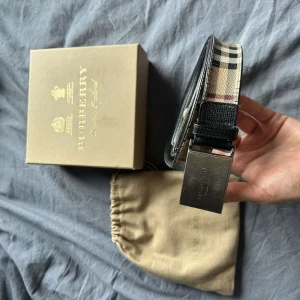Burberry rutigt skärp i skinn - Snyggt Burberry-bälte i klassiskt rutig design med beige, svart, vit och röd färg. Bältet har en bred rem i skinn och en robust metallspänne med Burberry-logga. Perfekt accessoar för att lyfta din outfit med en lyxig touch.