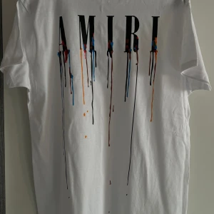 Vit AMIRI t-shirt med färgstänk - Vit t-shirt från AMIRI med svart logga framtill och coola färgstänk i blått, rött och orange som rinner ner från bokstäverna. Klassisk passform och rund hals. Perfekt för dig som gillar streetstyle och vill sticka ut.