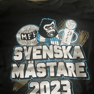 MFF Svenska Mästare 2023 t-shirt - Svart t-shirt med stort tryck på bröstet: MFF-logga, en gorilla med halsduk, pokal och texten 'Svenska Mästare 2023' i vitt och blått. Klassisk rund hals och korta ärmar. Perfekt för dig som vill hylla Malmö FF:s guldår.