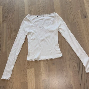 Vit långärmad topp med spets och rosett - Supersöt vit långärmad topp med ribbad struktur, spetskant runt den breda urringningen och en liten rosett framtill. Ärmsluten har vågig kant för extra detalj. Perfekt för dig som gillar en feminin och enkel stil.💗