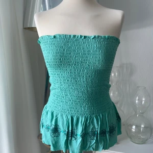 Grön bandeau tubtopp från italienska Terranova - Supersöt grön bandeau tubtopp från Terranova med smockad överdel och volangkant nertill. Toppen har knytning i ryggen och svarta blommiga broderier längs nederkanten. Perfekt för varma dagar och ger en riktigt somrig vibe. Du knyttet bak själv. 