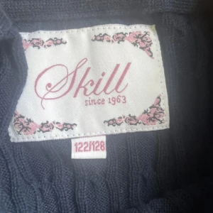 Mörkblå kabelstickad kofta från Skill - Mörkblå kabelstickad kofta från Skill med rund hals och knappar framtill. Långärmad och ribbade muddar vid ärmslut och nederkant. Perfekt för lager-på-lager och snygg till jeans eller kjol.