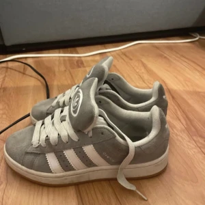Grå Adidas Campus sneakers i mocka - Snygga Adidas Campus sneakers i grå mocka med klassiska vita ränder på sidorna och vit logga på hälen och plösen. Skorna har rund tå, platt sula och vita snören. Perfekta för dig som gillar retrostil och streetwear.
