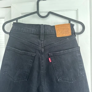 Svarta Levi's  ribcage crop flare jeans, hög midja - Säljer ett par svarta Levi's ribcage crop flare  jeans med hög midja och utsvängda ben. Klassisk femficksmodell med knappgylf och råa kanter nertill. Jeansen har den ikoniska läderpatchen bak och är tillverkade i kraftig denim. Perfekta för en cool och avslappnad stil.