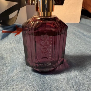 Hugo Boss The Scent Magnetic parfym - En lyxig parfymflaska från Hugo Boss, The Scent Magnetic. Flaskan är djupt lila med geometrisk design och har en elegant guldfärgad kork. Perfekt för dig som gillar exklusiva dofter och snygg design på parfymhyllan.
