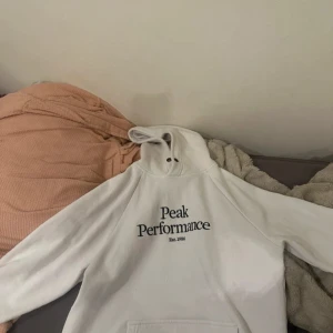 Vit hoodie från Peak Performance M - Vit hoodie från Peak Performance i storlek M. Klassisk modell med svart logga på bröstet, huva med snörning och stor magficka. Mjuk insida i fleece och långa ärmar. Perfekt för chill dagar eller när du vill ha en clean look.
