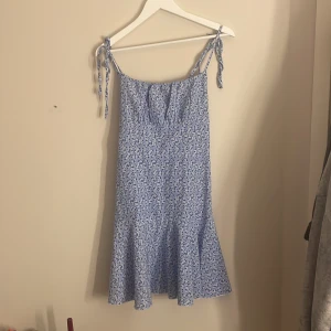 Blå blommig sommarklänning med knyt - Supersöt blå kortklänning med vita små blommor och knytbara axelband. Klänningen har en lätt utsvängd nederdel och romantisk vibe, perfekt för varma dagar. Tunn och luftig, med volangkant nedtill och smickrande passform.