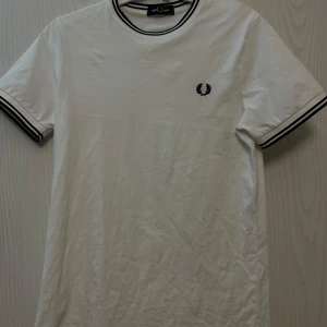 Vit Fred Perry t-shirt - Vit t-shirt från Fred Perry i bomull med svarta ränder på krage och ärmslut. Klassisk logga broderad på bröstet. Enkel och stilren modell med normal passform och korta ärmar. Perfekt för en clean look. Tröjan är köpt på Harrods i London och är helt äkta. Tröjan är i väldigt fint skick och är bara använd fåtal gånger. Skriv vid mer frågor eller funderingar🔥🔥