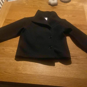 Svart kappa från Gina Tricot - Stilren svart kappa från Gina Tricot med klassisk krage och tre knappar framtill. Jackan har en rak, något croppad passform och är helfärgad utan mönster. Perfekt för dig som gillar en clean och tidlös look.
