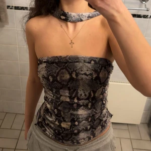 Ormmönstrad tubtopp från Zara - Säljer en snygg tubtopp från Zara med ormmönster i svart, grått och beige. Toppen är ärmlös och har en unik detalj med matchande choker runt halsen. Materialet är stretchigt och lätt, perfekt för att sticka ut med en edgy vibe.