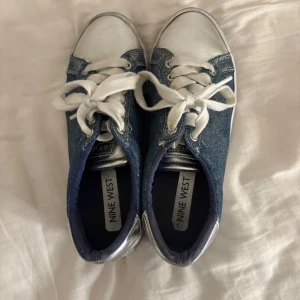 Blå denim sneakers från Nine West - Snygga sneakers från Nine West i blått denimtyg med vita detaljer och vita snören. Skorna har rund tå och platt sula, perfekt för en avslappnad och cool stil. Silverfärgade detaljer vid hälen ger extra edge.