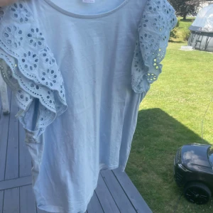 Ljusblå topp med volangärmar - Supersöt ljusblå topp med broderade volangärmar i bomull. Ärmarna har ett snyggt hålmönster och ger en romantisk vibe. Perfekt för dig som gillar detaljer och vill sticka ut lite extra. Rund halsringning och mjukt material. En liten choklad flek men inte andvönd mer än 3 gånger för dig som är bra på att ta bort flekar men även andra jag är inte så bra😹