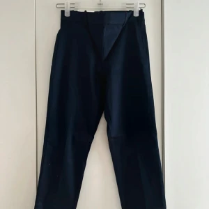 Mörkblå byxa/slacks med regular waist - Snygga mörkblå ankelbyxor med klassisk rak passform Byxorna har hällor för bälte, diskreta bakfickor och är tillverkade i ett slätt tyg som ger en stilren look. Perfekta för dig som gillar en clean och dressad stil. Dragkedjan är på sidan