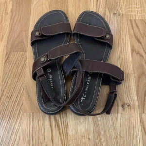Bruna sandaler från Tamaris i skinn - Snygga bruna sandaler från Tamaris med breda remmar i skinn och vita kontrastsömmar. Justerbara remmar med metallknappar och en robust svart sula ger både stil och komfort. Perfekta för varma dagar och enkel att matcha med sommarens outfits.Pris kan diskuteras !                                              OBS! Köparen står för frakt!