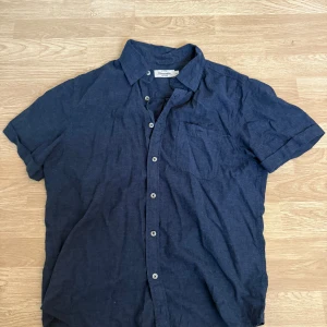 Mörkblå linneskjorta Dressmann M - Snygg mörkblå skjorta från Dressmann i linnemix. Skjortan har korta ärmar, klassisk krage, bröstficka och knäppning framtill. Perfekt för varma dagar när du vill vara både chill och stilren.