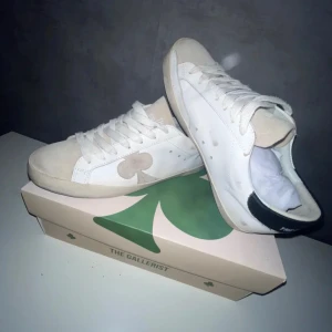 Vita sneakers från The Gallerist - Snygga vita sneakers från The Gallerist med beige mockadetaljer och en stor klöver på sidan. Skorna har svart hälparti, rund tå och klassisk snörning. Perfekta för dig som gillar stilrena och trendiga sneakers med unika detaljer.