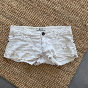 Vita jeansshorts från Bershka - Snygga vita jeansshorts från Bershka med låg midja, slitna detaljer framtill och råa kanter. Klassisk femficksmodell med knappar på bakfickorna och cool streetkänsla. Perfekta för varma dagar och en avslappnad stil.