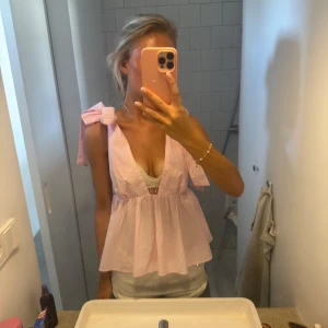 Ljusrosa blus med rosetter på axlarna - Supersöt ljusrosa blus med djup v-ringning och stora rosetter på axlarna. Blusen är ärmlös och har en volangig, luftig passform som ger en romantisk vibe. Perfekt för varma dagar och somriga outfits.