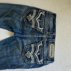 Blå Bootcut jeans från Rock Revival - Snygga blå jeans från Rock Revival med bootcut passform. Jeansen har markerade slitningar, kontrastsömmar och detaljerade bakfickor med broderi och nitar. Klassisk femficksmodell i jeansmaterial med coola detaljer och logga i midjan. PRIS DISKUTERAS 