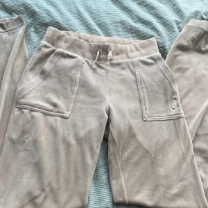 Beige Juicy Couture - Supermjuka beige mjukisbyxor från Juicy Couture med bred resår och snörning i midjan. Byxorna har stora fickor fram och bak med lock och knappar, samt broderad JC-logga på ena fickan. Byxorna har några få små defekter som går att putsa om bort därav priset. Perfekta för en chill och trendig look.💗