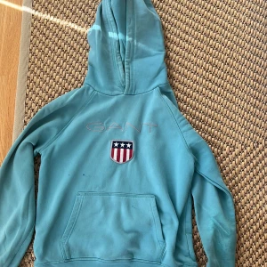 Turkos hoodie från Gant med logga - Turkos hoodie från Gant med broderad logga och sköld på bröstet. Klassisk känguruficka framtill och stor huva. Perfekt för en chill och sportig look. Mjuk bomullskänsla och långa ärmar.