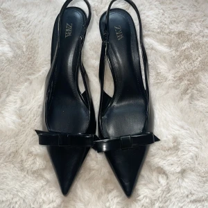 Svarta pumps med rosett från Zara - Stilrena svarta pumps från Zara med spetsig tå och elegant rosettdetalj framtill. Skorna har öppen häl med smal rem och är tillverkade i glansigt syntetmaterial. Endast använda en gång