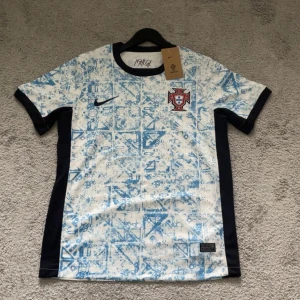 Portugal Nike fotbollströja blåvit - Portugal fotbollströja från Nike med unikt blått mönster på vit bas. Tröjan har svarta detaljer längs sidorna och runt ärmslut samt krage. Broderad Portugal-logga på bröstet och Nike Swoosh. Tillverkad i lätt och ventilerande material, perfekt för match eller träning.