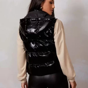 Svart glansig dunväst med huva - Svart, glansig dunväst med huva och dragkedja framtill. Västen har en quiltad look och breda kanaler, vilket ger en cool och sportig vibe. Perfekt att slänga över en hoodie för en trendig streetstyle. Några år gammal och använd mycket, snörena finns inte kvar. 