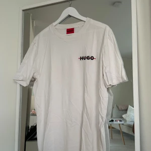 Vit t-shirt från Hugo med logga - Vit t-shirt från Hugo med klassisk rund halsringning och svart logga med röd detalj på bröstet. Enkel och stilren design, perfekt till jeans eller shorts. Tillverkad i mjuk bomull för en skön känsla hela dagen.