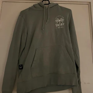 Grön hoodie från Vacay Dope Co - Snygg grön hoodie från Vacay Dope Co med tryck av palm och text både fram och bak. Tröjan har huva med snörning och en stor magficka. 